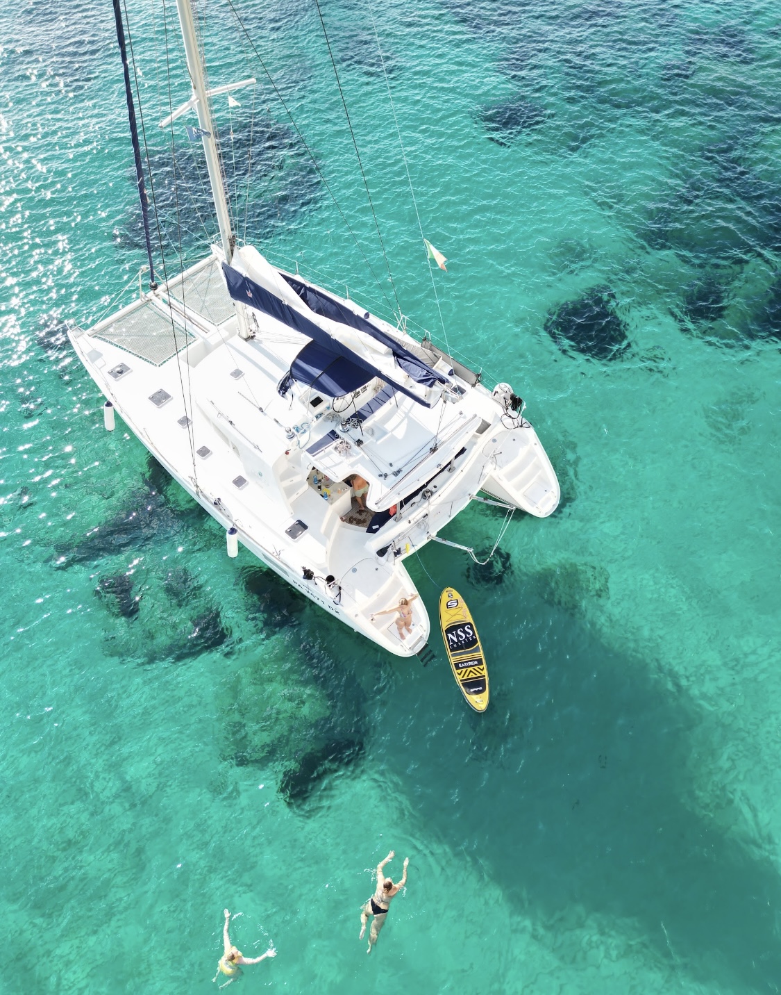 Aerial view of a white catamaran anchored over clear turquoise water, with people swimming beside it and a yellow paddleboard towed behind