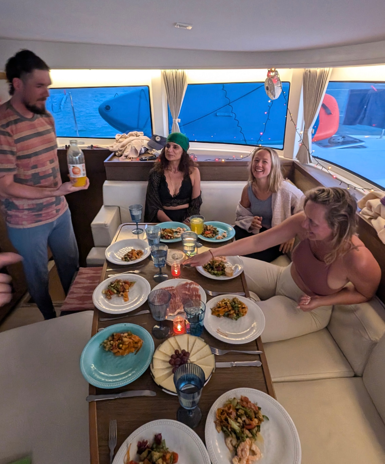 Travellers sharing a candlelit tapas dinner in the yacht's salon at dusk, fairy lights above and blue water beyond the windows