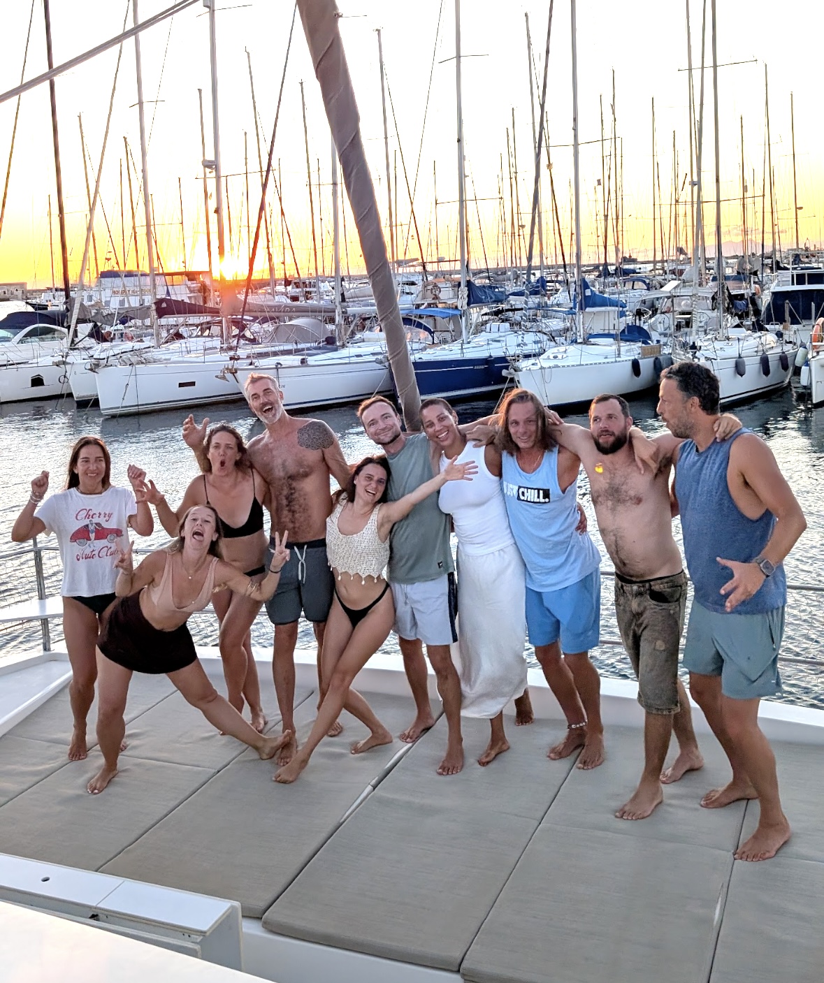 Group of travellers posing together on the deck of a catamaran at sunset, marina masts behind them